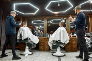 Two barbers holding mirrors and showing haircuts to father and son sitting in armchair at modern barbershop. Family, father and son visiting barber shop. Child in barbershop. Mens haircut