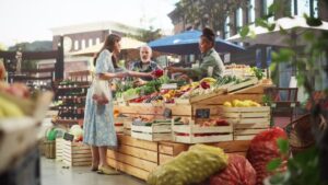 Middle Aged Diverse Couple Running a Small Business on a Farmers Market, Selling Organic Fruits and Vegetables. Young Female Shopping For Food, Choosing a Fresh Courgette or Zucchini