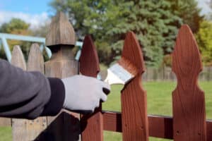Man is painting wooden fence with brown paint outside