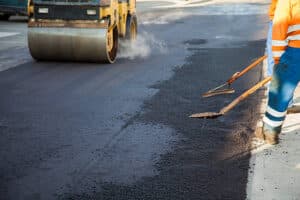 Two manual workers helping to level the asphalt surface for a steamroller to press.